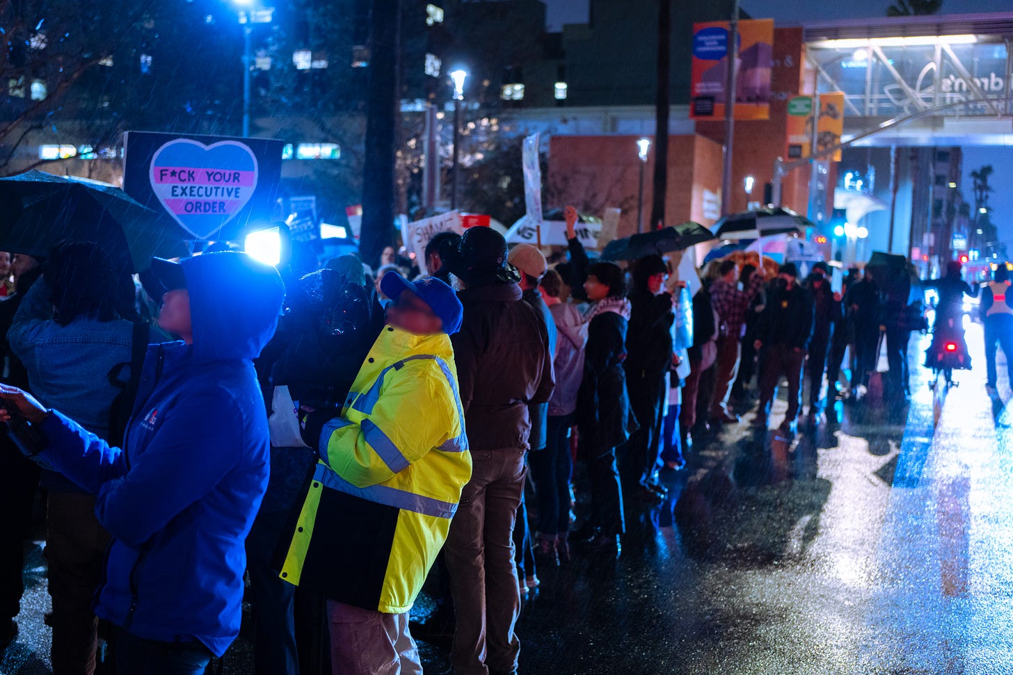 A line of people on the edge of the protest hold signs and umbrellas, with the CHLA pedestrian bridge visible in the background