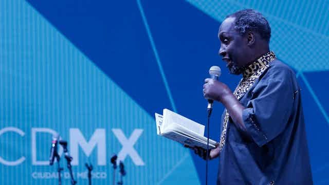 An older African man dressed in blue traditional attire on a stage against a blue backdrop. He holds a book in his one hand and a microphone in the other. An older African man dressed in blue traditional attire on a stage against a blue backdrop. He holds a book in his one hand and a microphone in the other.