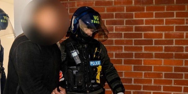 Police officers escorting a person from a building at night. The man's face is pixelated to hide his identity.