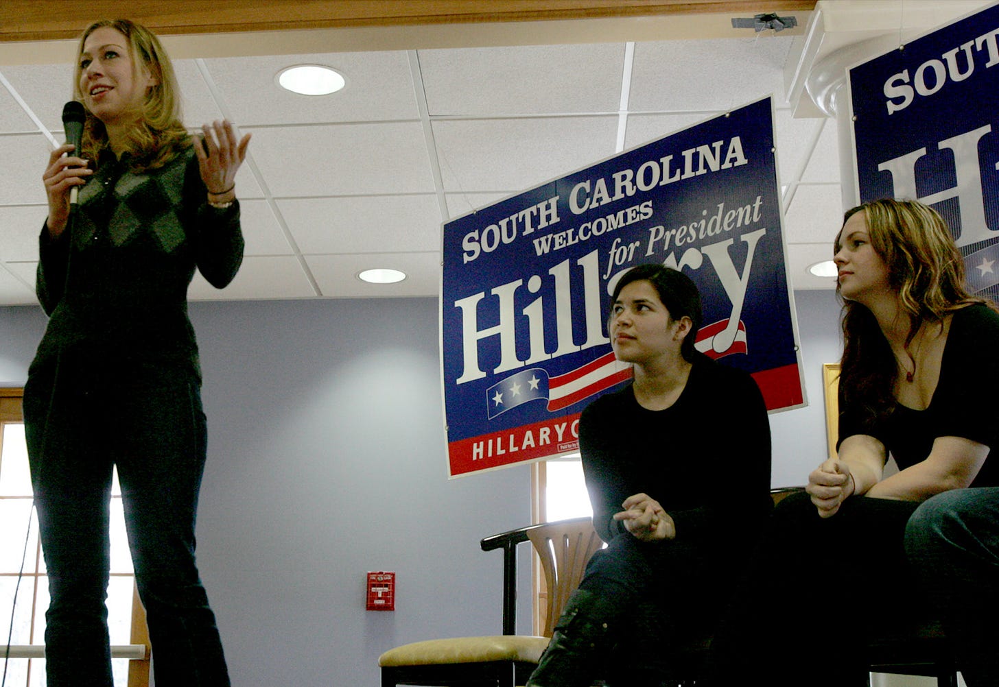 America Ferrera and Amber Tamblyn sit behind Chelsea Clinton as she speaks into a microphone. Behind them, large signs read “South Carolina welcomes Hillary for President”