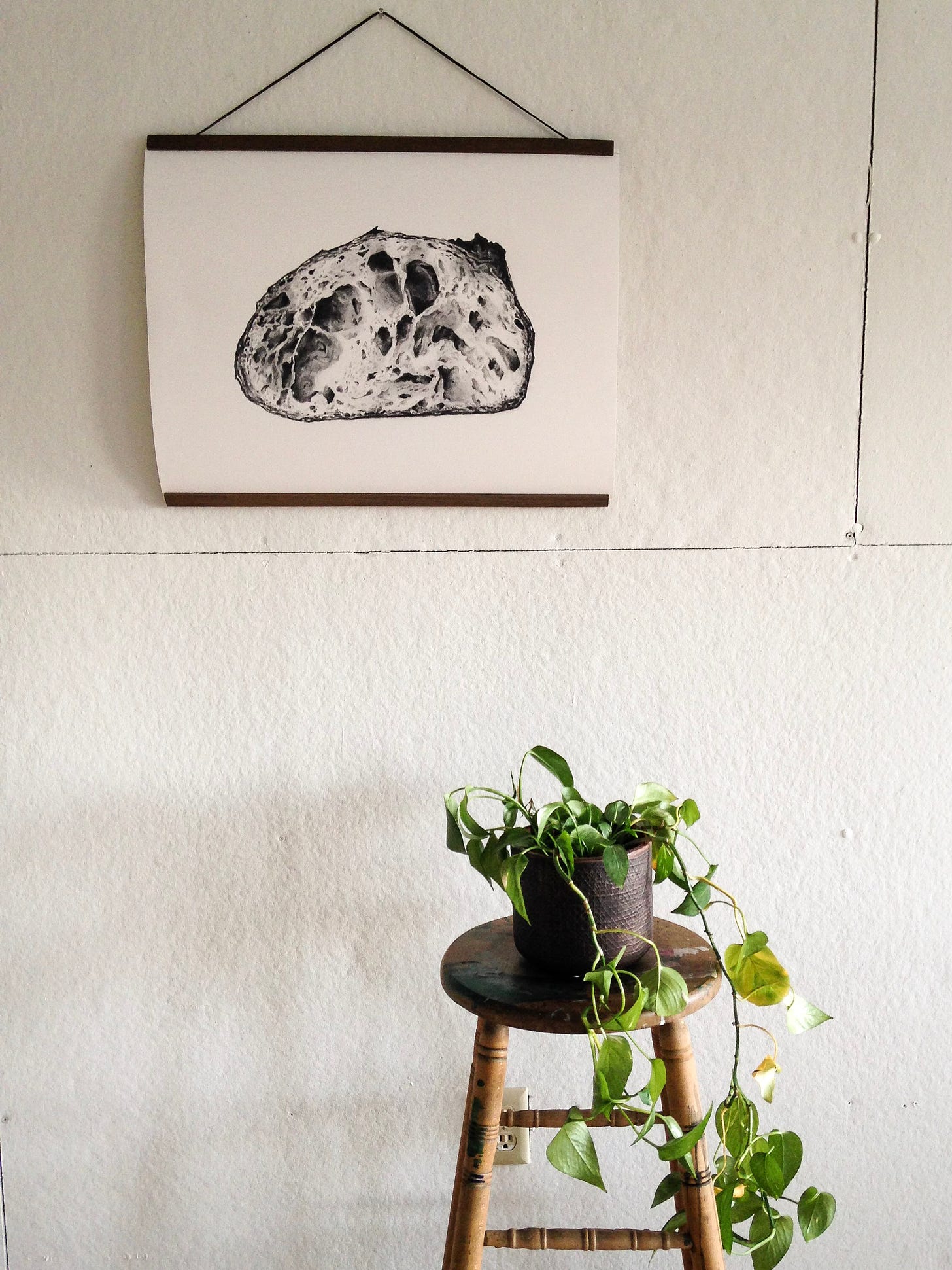 A large sourdough print hanging on a white wall, with a green plant on a stool below it. 