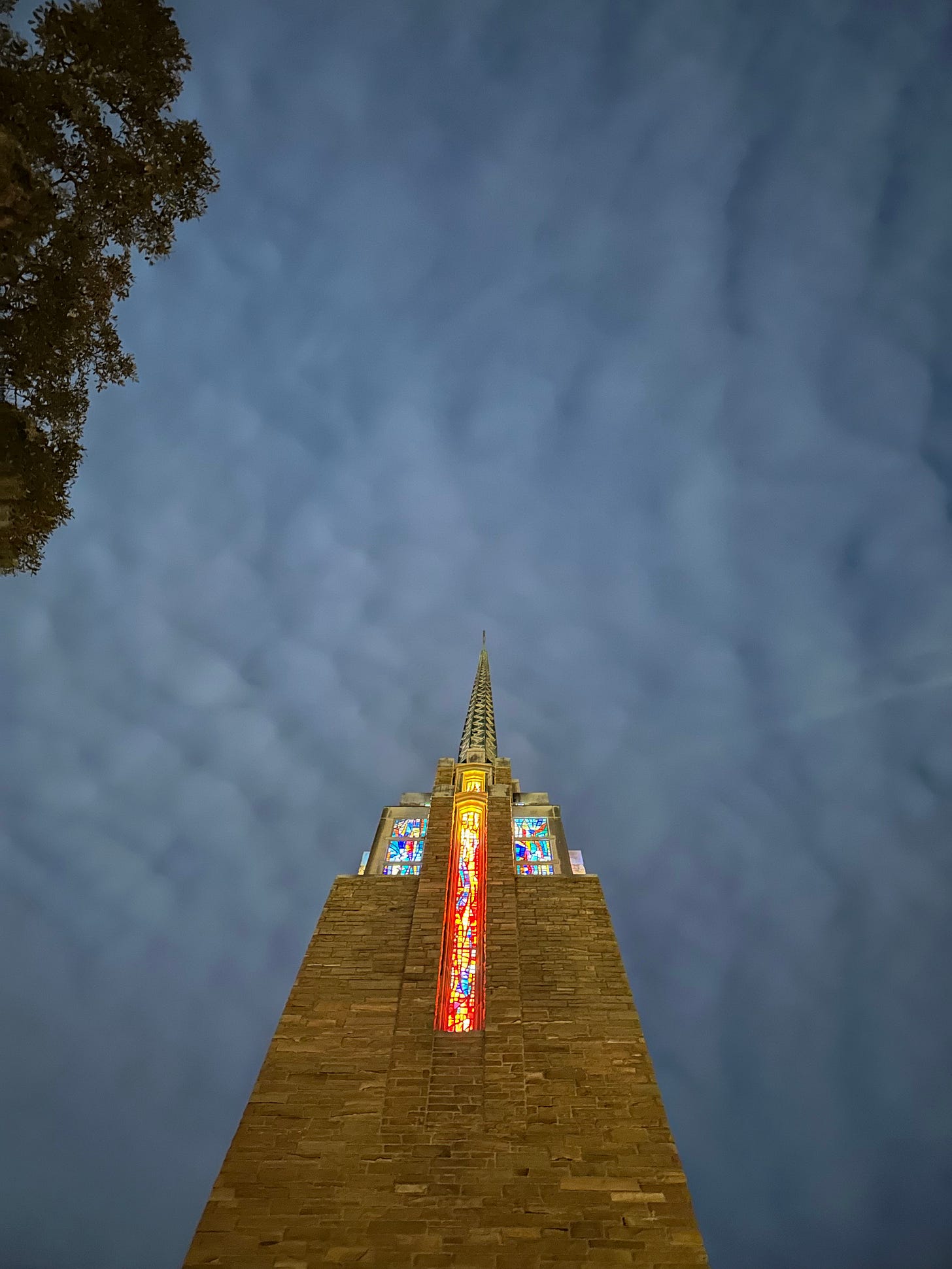 Looking up along a brick church steeple with lit stained glass against a cloudy sky
