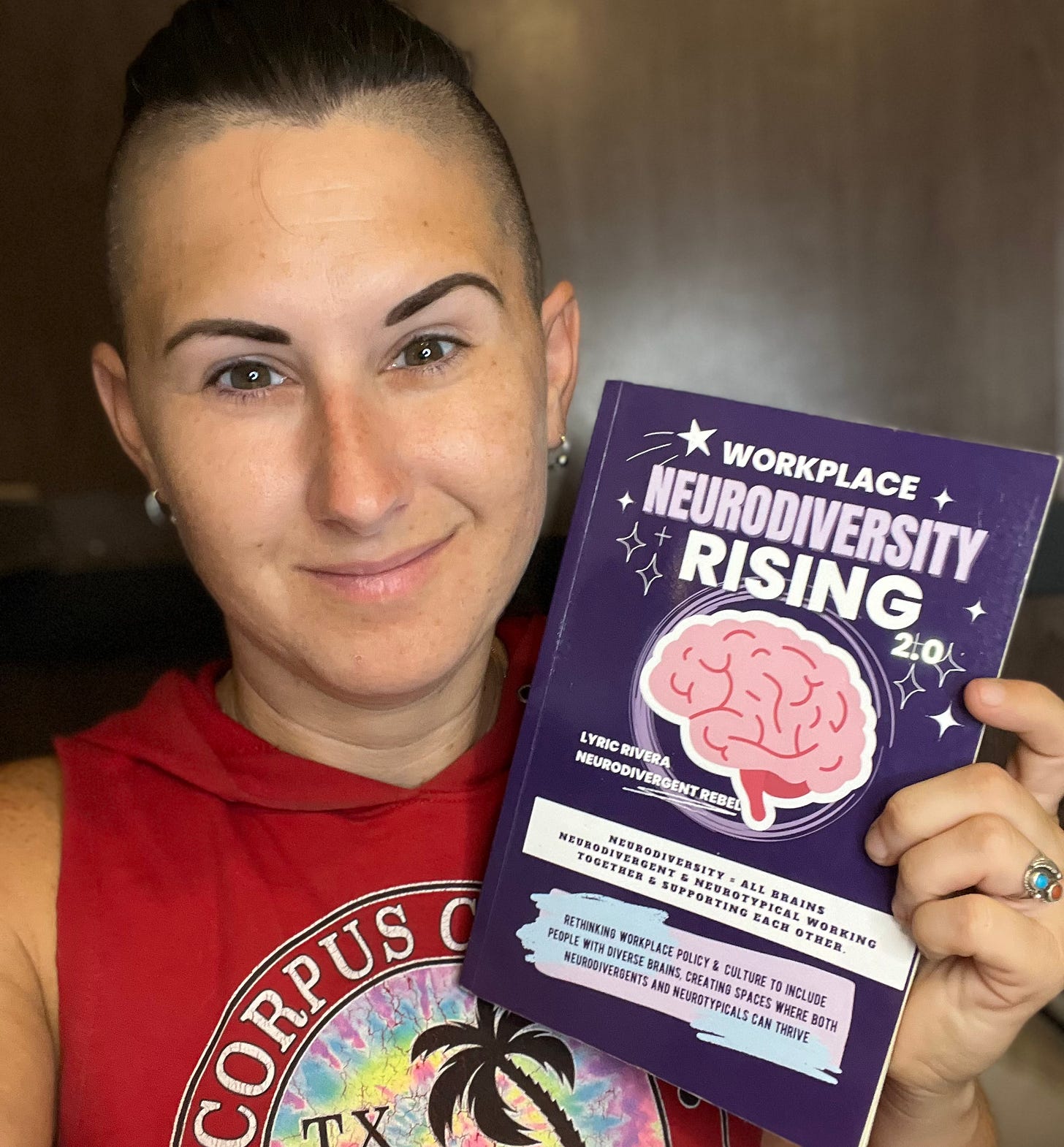 Lyric Rivera holding up a copy of their book, Workplace NeuroDiversity Rising and smiling at the camera, posing happily with their book.