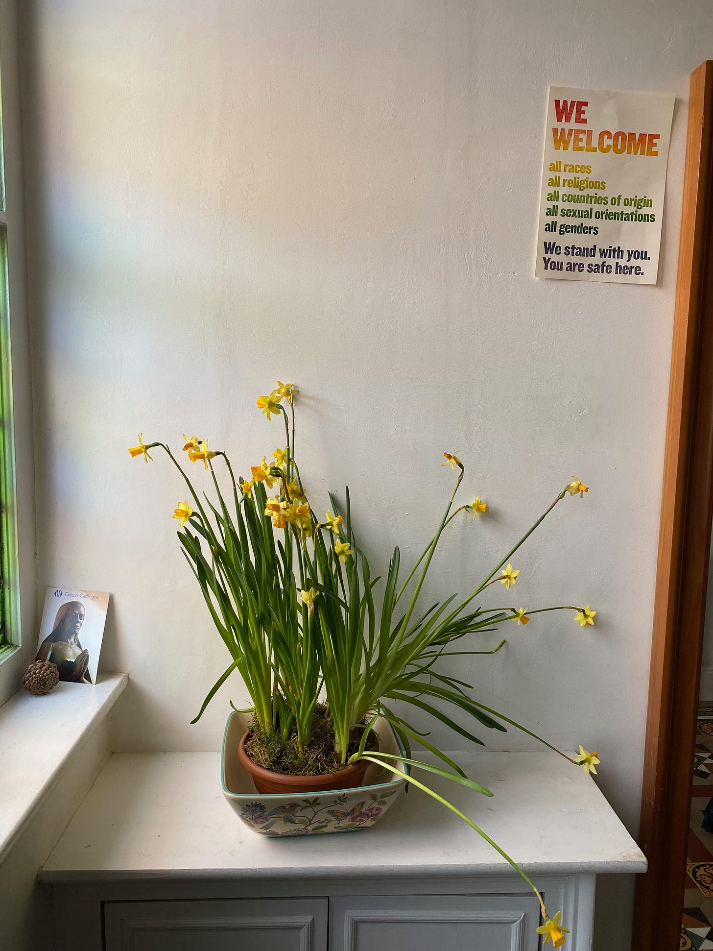 A bowl of daffodils begin to fade in a ceramic pot on a shelf in an interior room against a white wall