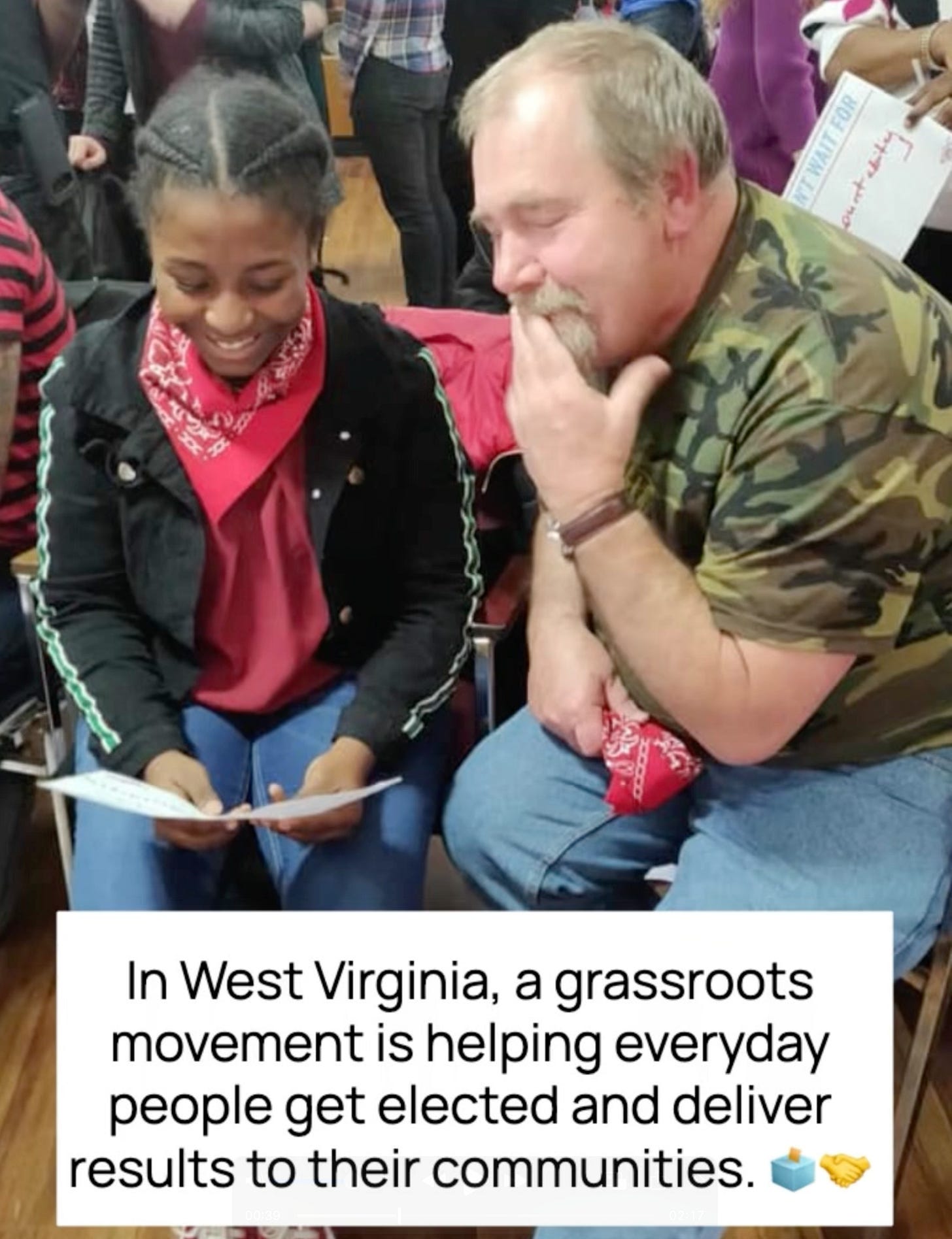 A young Black woman and an older white man sit closely together, smiling and looking down at a sheet of paper she’s holding. They’re both wearing red bandanas, suggesting solidarity or shared purpose. The setting appears to be a community gathering with others in the background. Overlaid text reads: “In West Virginia, a grassroots movement is helping everyday people get elected and deliver results to their communities.” Emoji symbols of a ballot box and handshake accompany the message.