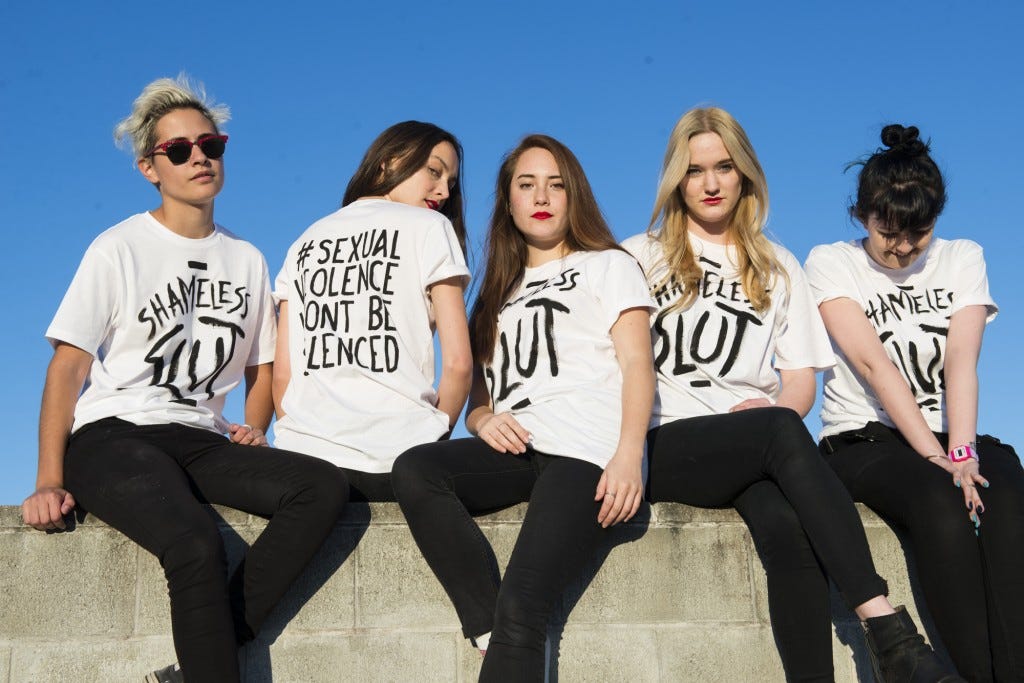 5 women sitting on a wall looking at the camera, their shirts say 'Sexual violence won't be silenced' on the back and 'shameless slut' on the front