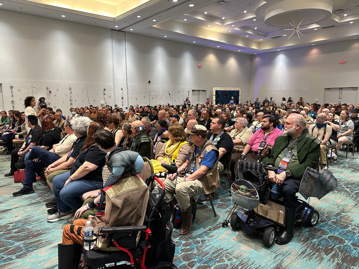 An absolutely packed convention room, many attendees in Star Wars costumes, with speaker Mikhael Tara Garver moving among them