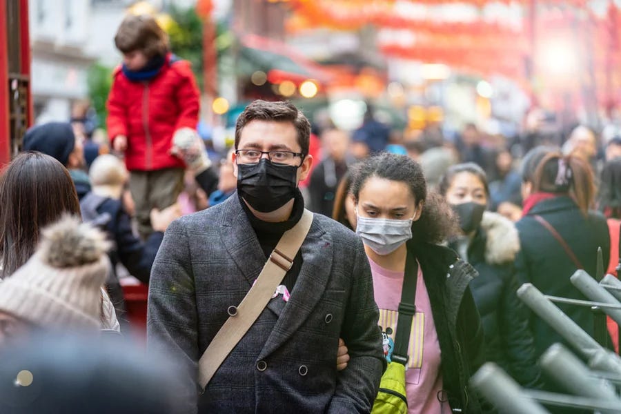 Young couple wearing protective face masks walking through a crowded urban street during flu season. The man wears a black mask and gray coat while the woman wears a surgical mask and pink sweater, surrounded by a mix of masked and unmasked people in a busy public space. Young couple wearing protective face masks walking through a crowded urban street during flu season. The man wears a black mask and gray coat while the woman wears a surgical mask and pink sweater, surrounded by a mix of masked and unmasked people in a busy public space.
