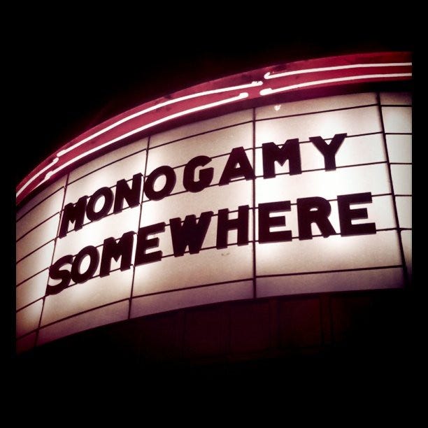 This is a dark photo -- and on some sort of movie marquee sign it says, "Monogamy Somewhere" in block all caps. red neon surrounding it