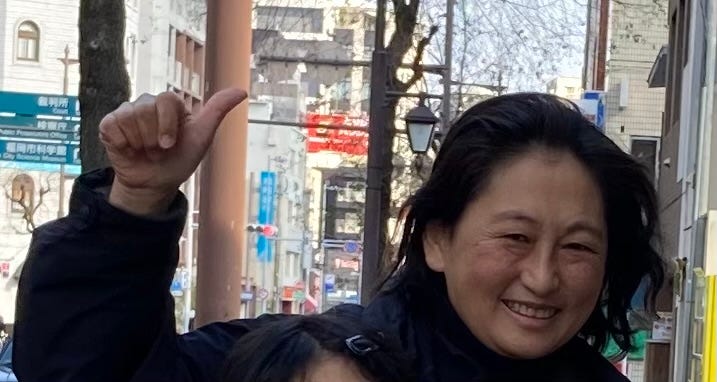 a Japanese woman smiles and gives a thumbs-up in an urban setting