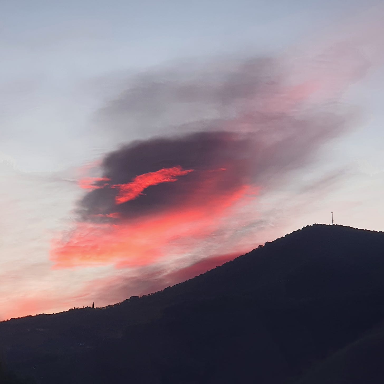 Ragged lenticular cloud mass in hues of brilliant pink and dark violet over low hills, lit from below by the rising sun.