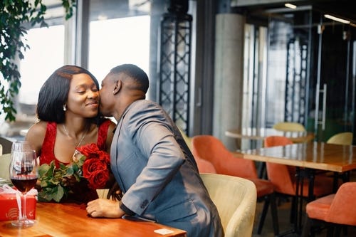 Young couple on date night at a fine dining restaurant. He is kissing her cheek while she holds a bouquet of red roses and there are two glasses of red wine and a gift on the table.