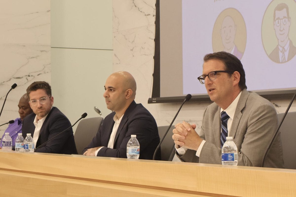 Four District 5 candidates seated at a panel discussion table in a conference room; three men in suits are visible in the foreground.