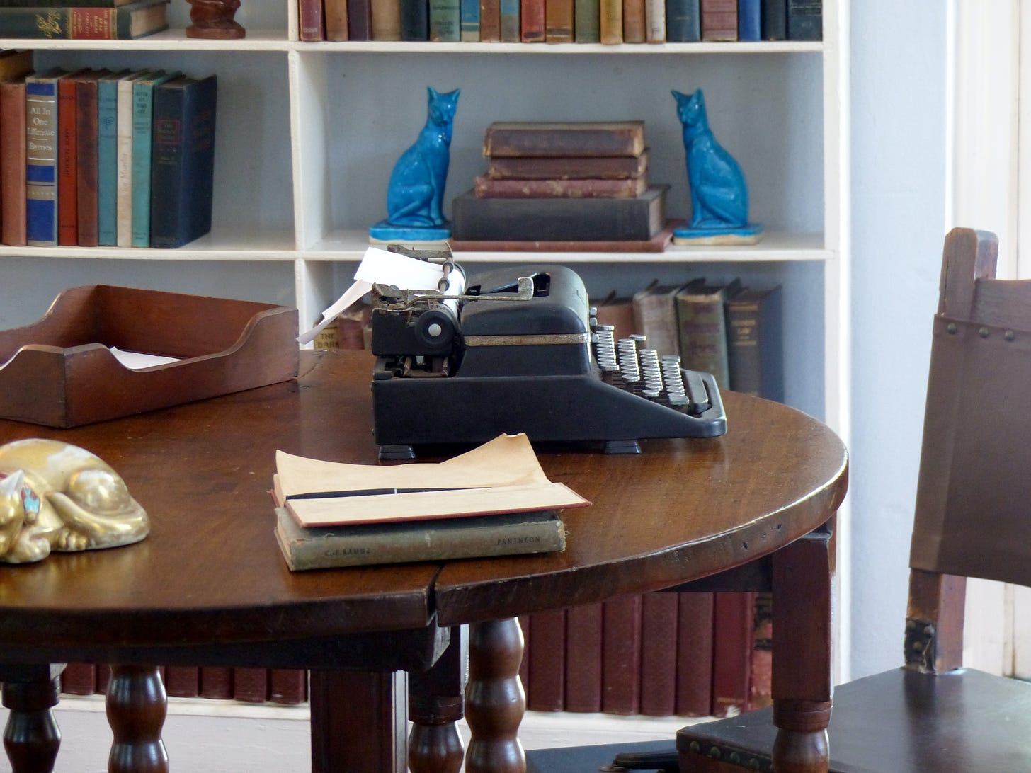 A black tyepwriter sits on a brown circular desk. Behind it is a bookshelf with books and two blue cat figurines.