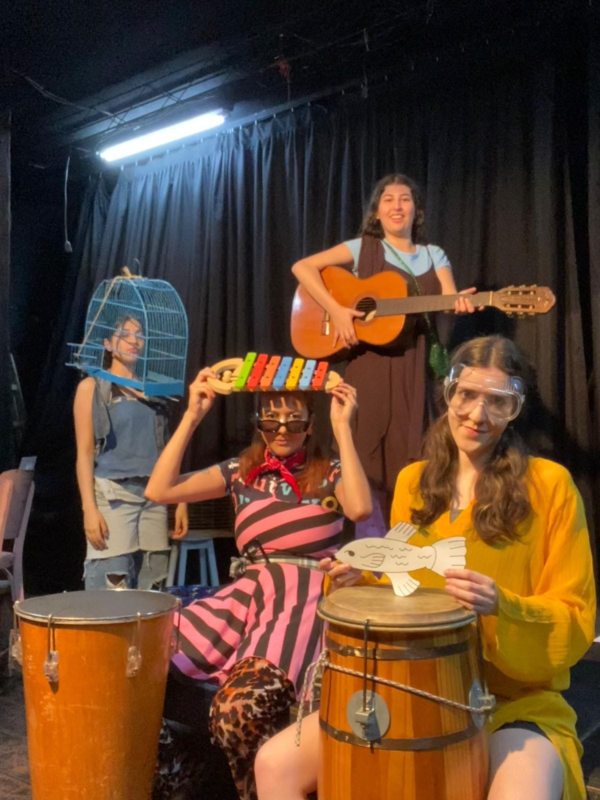 Foto de 4 atrizes. Da esquerda para a direita, A primeira; mais ao fundo está vestida toda de jeans com uma gaiola na cabeça; a segunda mais à frente está de listras, óculos escuros e um xilofone apoiado na cabeça, ao fundo, bem atrás uma outra está de vestido marrom segurando um violão bem no alto, e mais à direita estou eu, de amarelo, óculos epi transparente, uma sardinha de papel nas mãos e um atabaque na frente. Estamos em um palco com cortinas pretas atrás