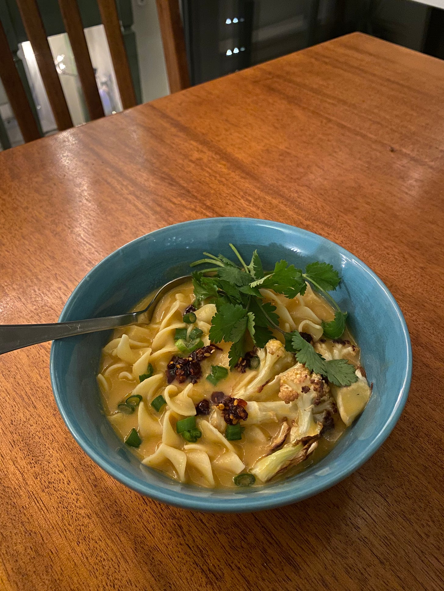 A blue bowl of the soup described above. Curly egg noodles in a creamy orange broth, topped with cilantro, roasted cauliflower, an chili crisp. There's a spoon sticking out at the back of the bowl.