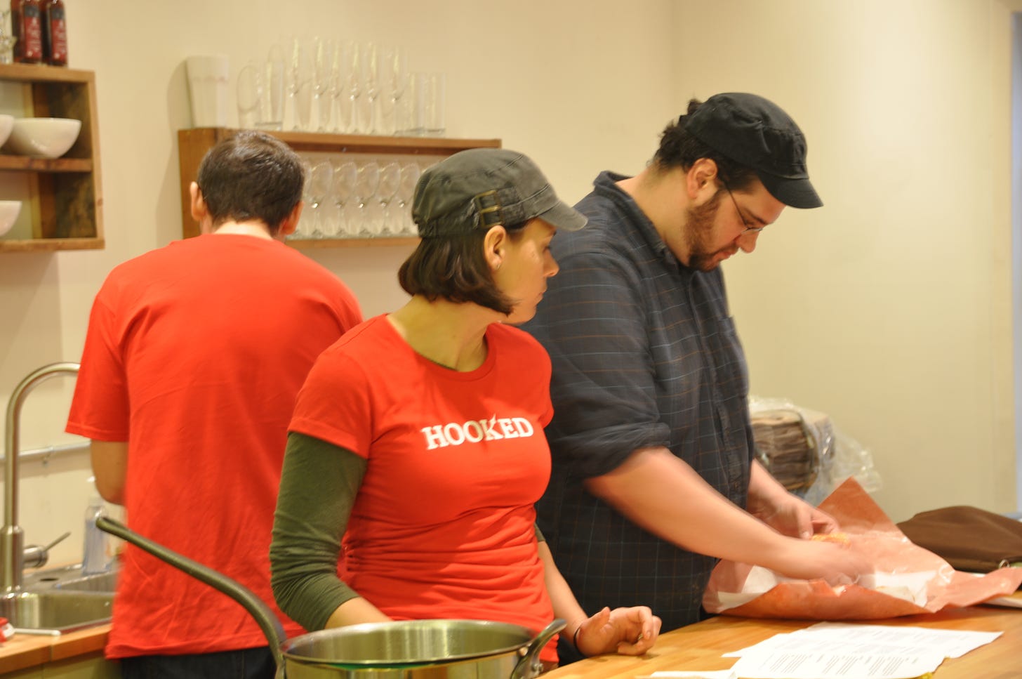 three people standing in a room, two people face a counter where they are looking at something inside of butcher paper, a pan is in the foreground.