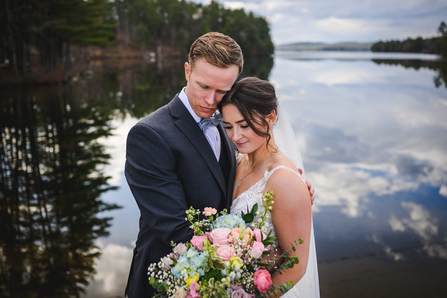 A bride and groom looking down in front of a picturesque lake with clouds reflecred in the water