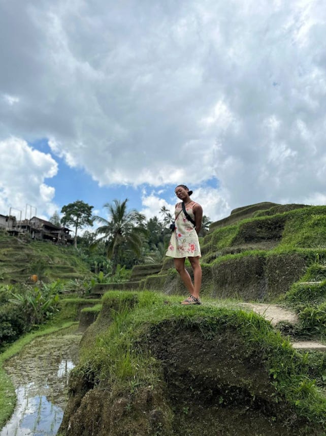 Nathalie stands on a hill of a rice field. She is wearing a yellow and pink dress and lean her head slightly to the left. She has her arms resting against her back