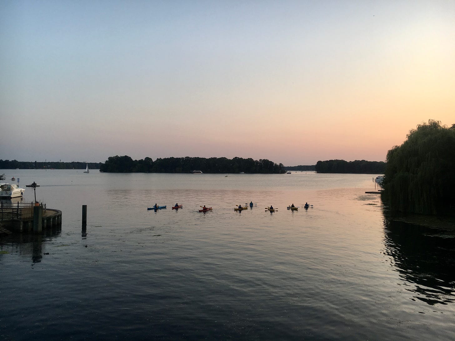 Ein See im Abendlicht, im Vordergrund eine Gruppe Kajakfahrer*innen, im Hintergrund eine Insel und ein Segelboot.