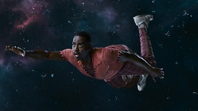 A man in an orange v-neck polo shirt and check trousers floats through space reaching towards something just out of shot