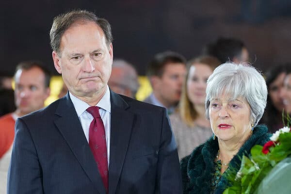 Justice Samuel A. Alito Jr., wearing a dark suit and red ties, stands next to his wife, Martha-Ann Alito, who is wearing a green coat.