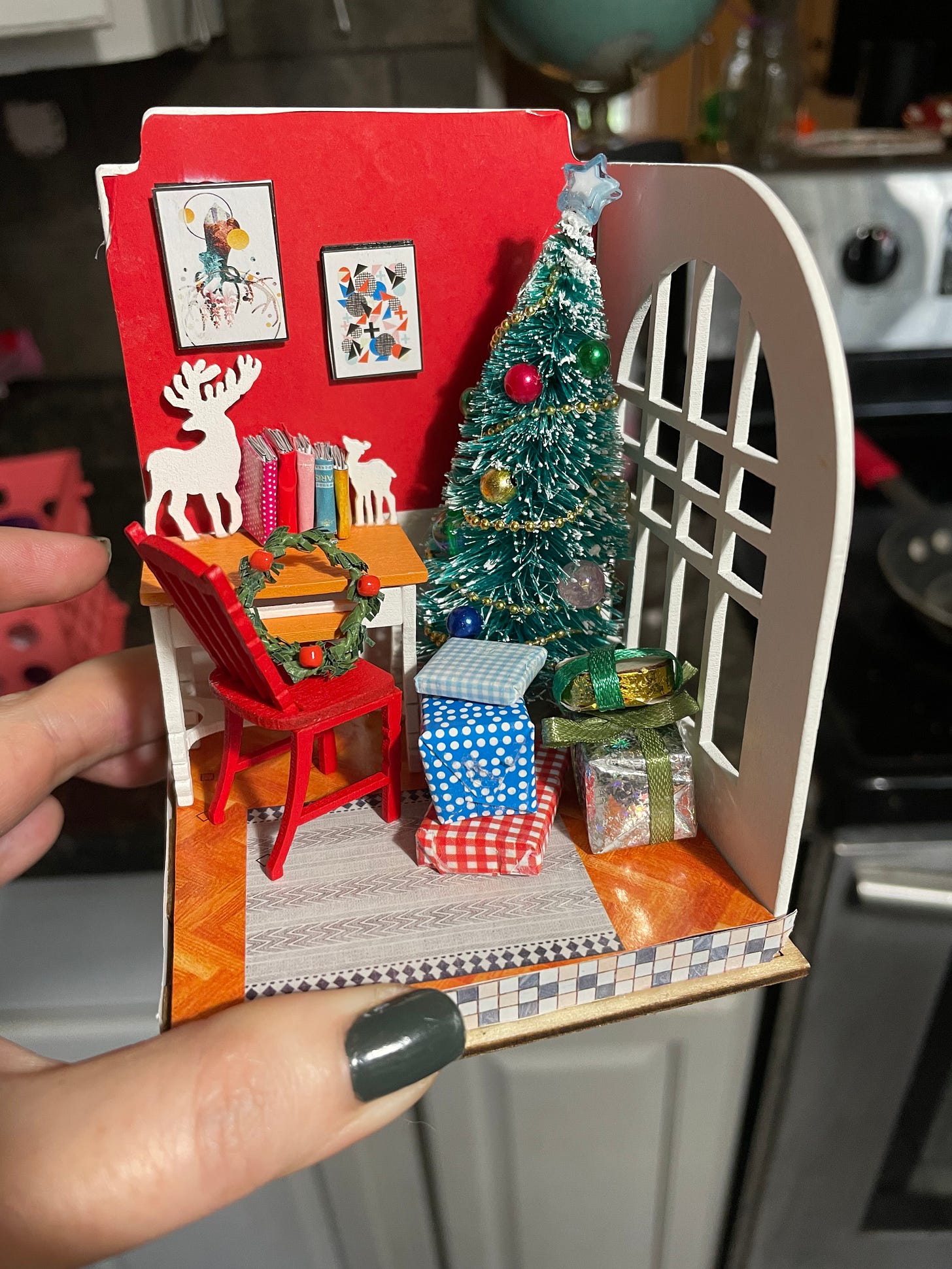 a tiny miniatures set with a red chair, a christmas tree, and paintings up on the walls. There is a tiny desk with tiny books and a few shiny wrapped presents. 