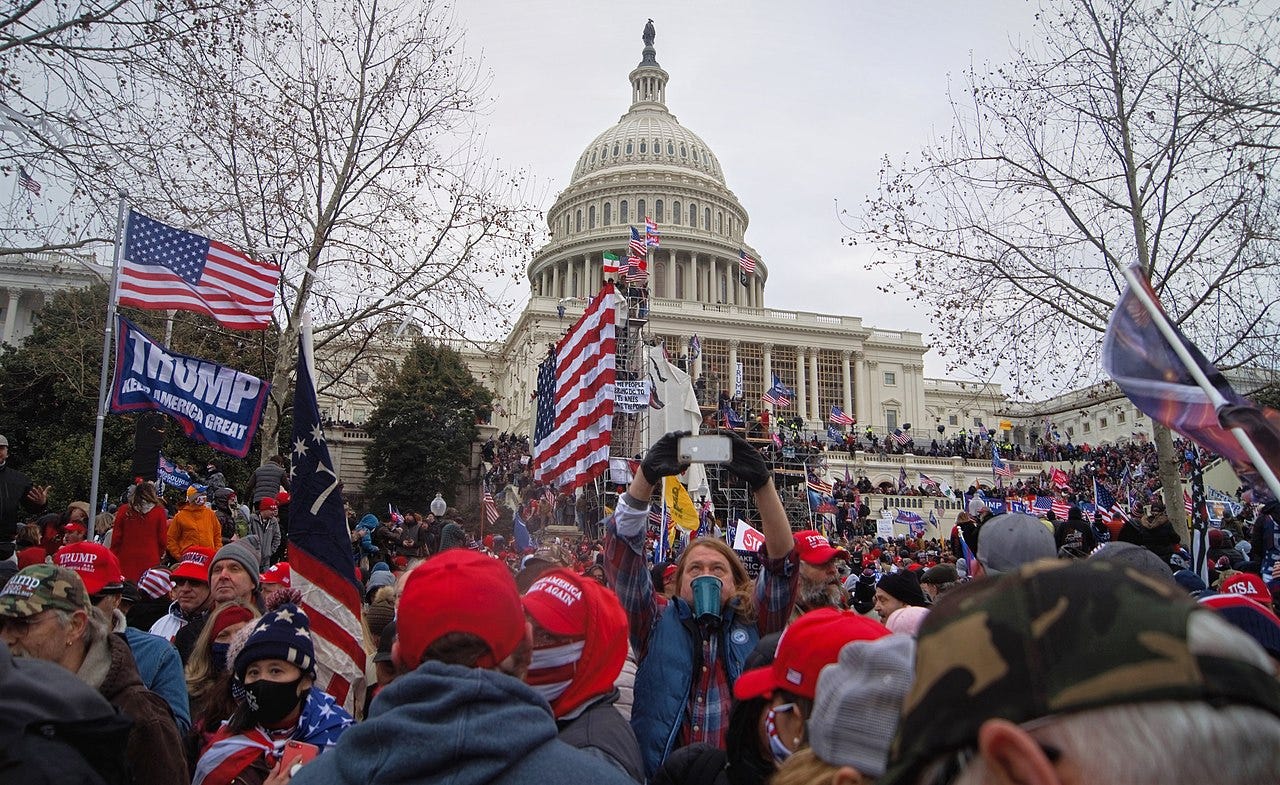 A crowd carrying Trump banners and American flags surrounding the US Capitol building. A man in the center of the picture is taking a selfie of himself with the Capitol in the background.