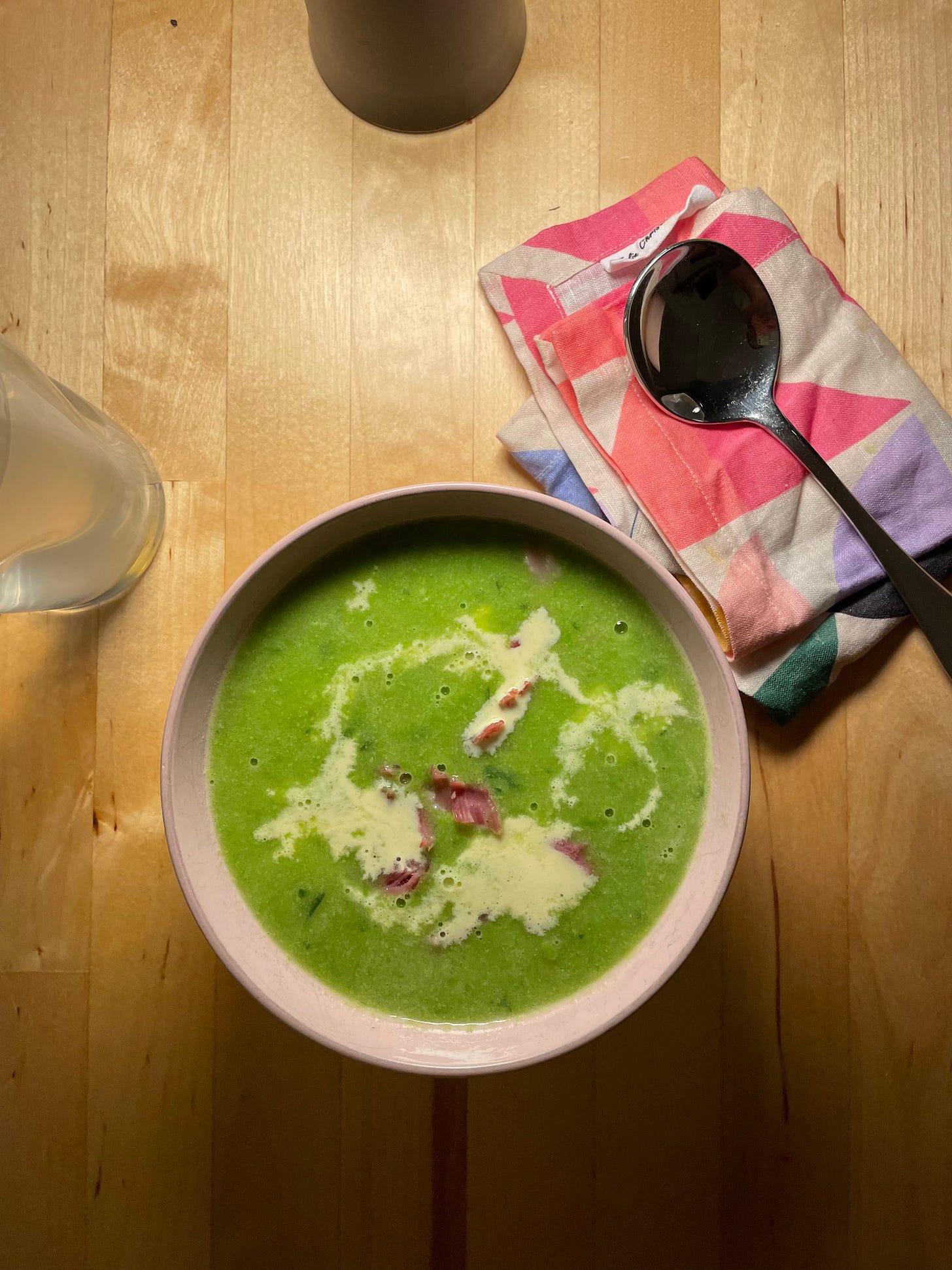 Pink bowl featuring bright green pea and ham hock soup, made with frozen peas. Chunks of ham hock are in the bowl and it's finished with a drizzle of cream. A patterned napkin and glass of coconut water are nearby. The table is lit by a small mushroom lamp.