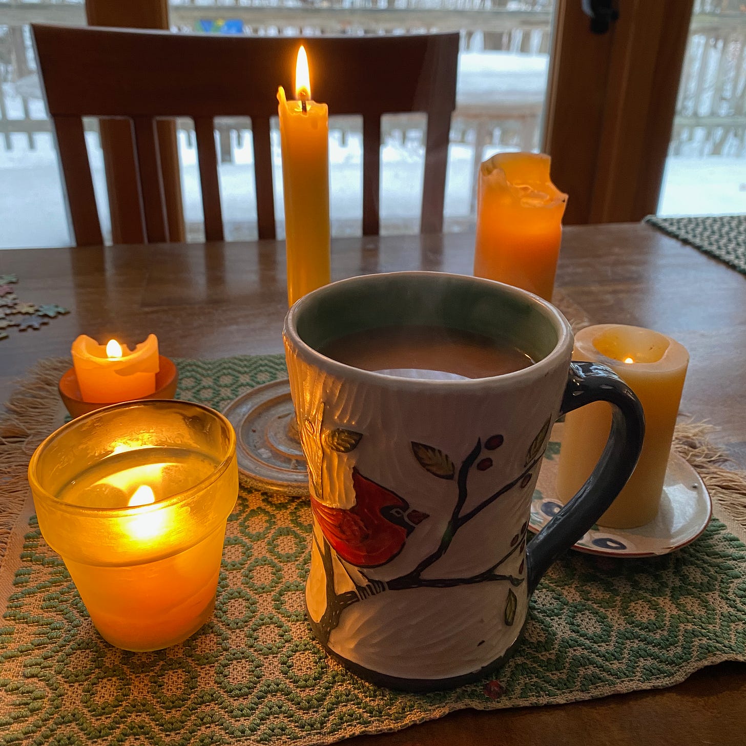 A ceramic mug of tea with a cardinal painted on it surrounded by several lit candles of various sizes.