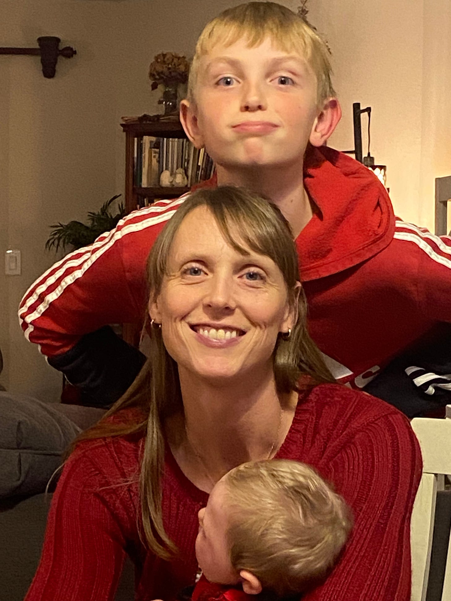 A smiling mother in red sits at home with her young son standing behind her and a baby in her arms. The image reflects a story of resilient faith through loss, maternal love, and the healing power of Jesus Christ during a year of trials, personal growth, and spiritual renewal.