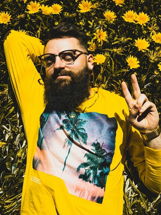 Man with beard in a yellow t-shirt lying on hte grass showing a V- Sign.