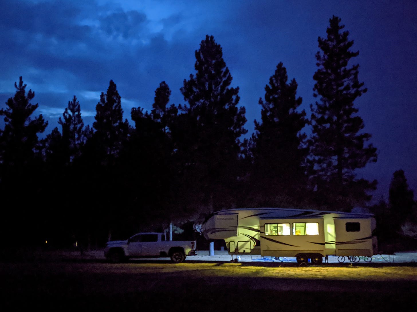 A pick-up truck and fifth wheel travel trailer, lit from within with a warm glow, set against a background of tall pine trees which are silhouetted against a dark blue night sky