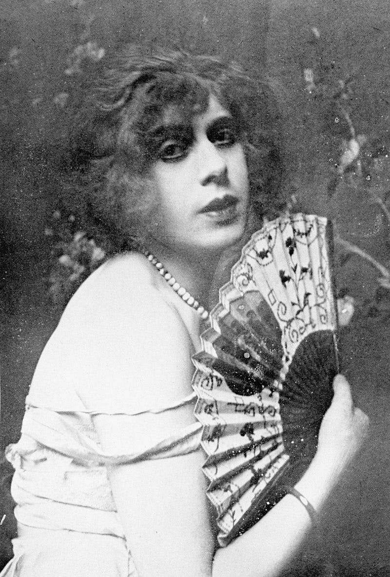 Black-and-white photograph of Lili Elbe. She is a white woman with dark hair. She is wearing a sunflower dress and jewelry and holding a hand fan.