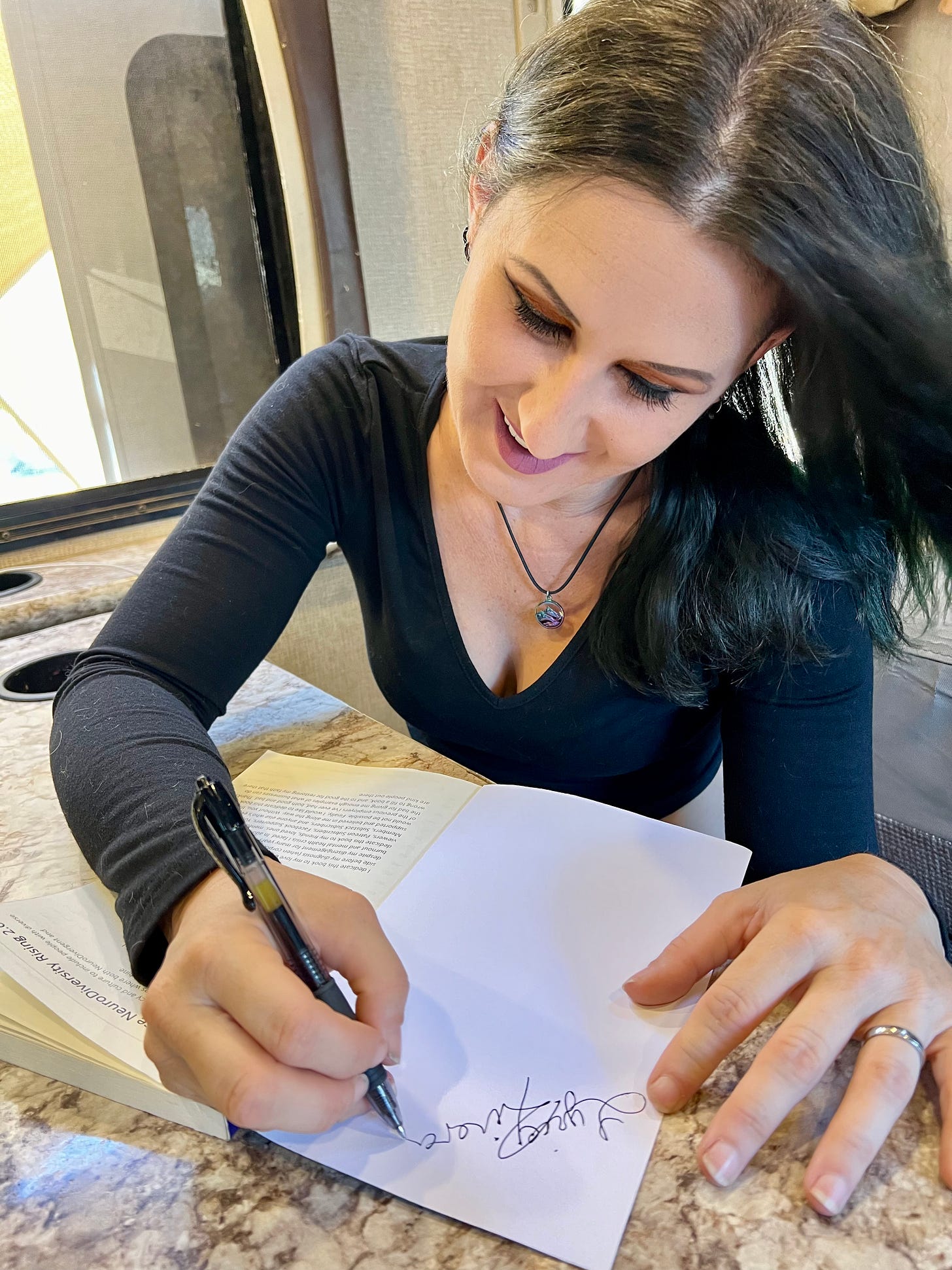 Image of Lyric, sitting at their RV dining room table signing a book.