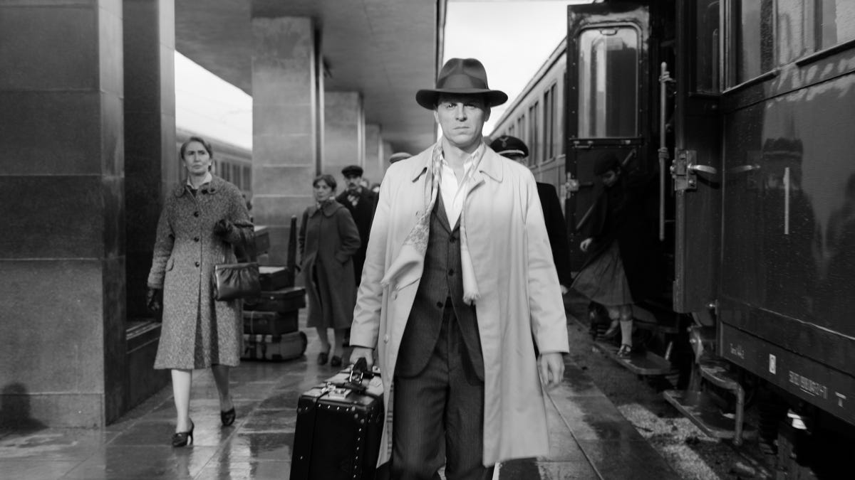 A man in a hat and coat carries a suitcase as he walks next to a train.