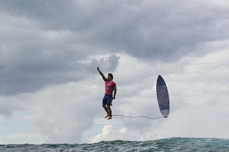 A surfer in a pink jersey appears to be levitating above the ocean surface. His right arm is raised with index finger pointing skyward in a triumphant pose. His surfboard, still attached by a leash, mirrors his posture perfectly beneath him. The scene is set against a dramatic cloudy sky and turbulent sea.