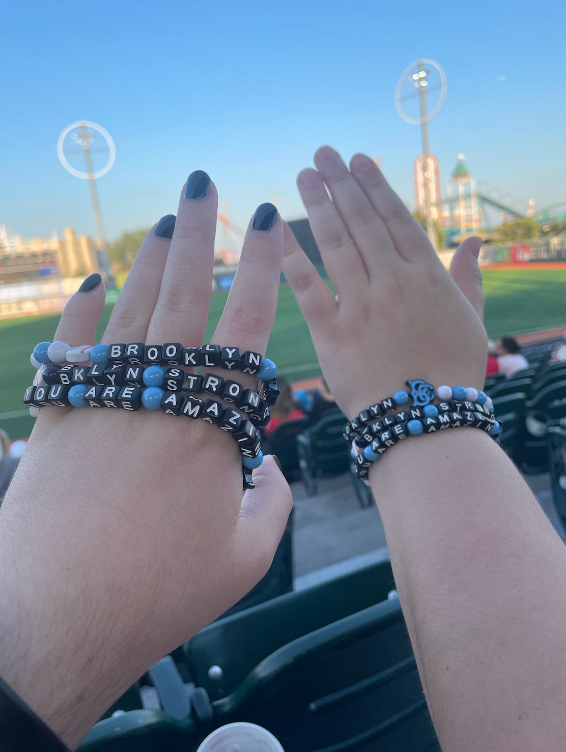 two hands in front of a baseball field, holding up beaded friendship bracelets