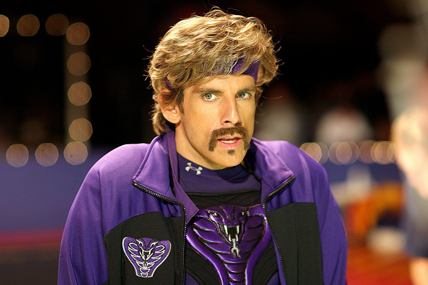 a man with floppy blonde hair and a mustache dressed in all purple looks at the camera in a suspicious way. it's ben stiller jsyk