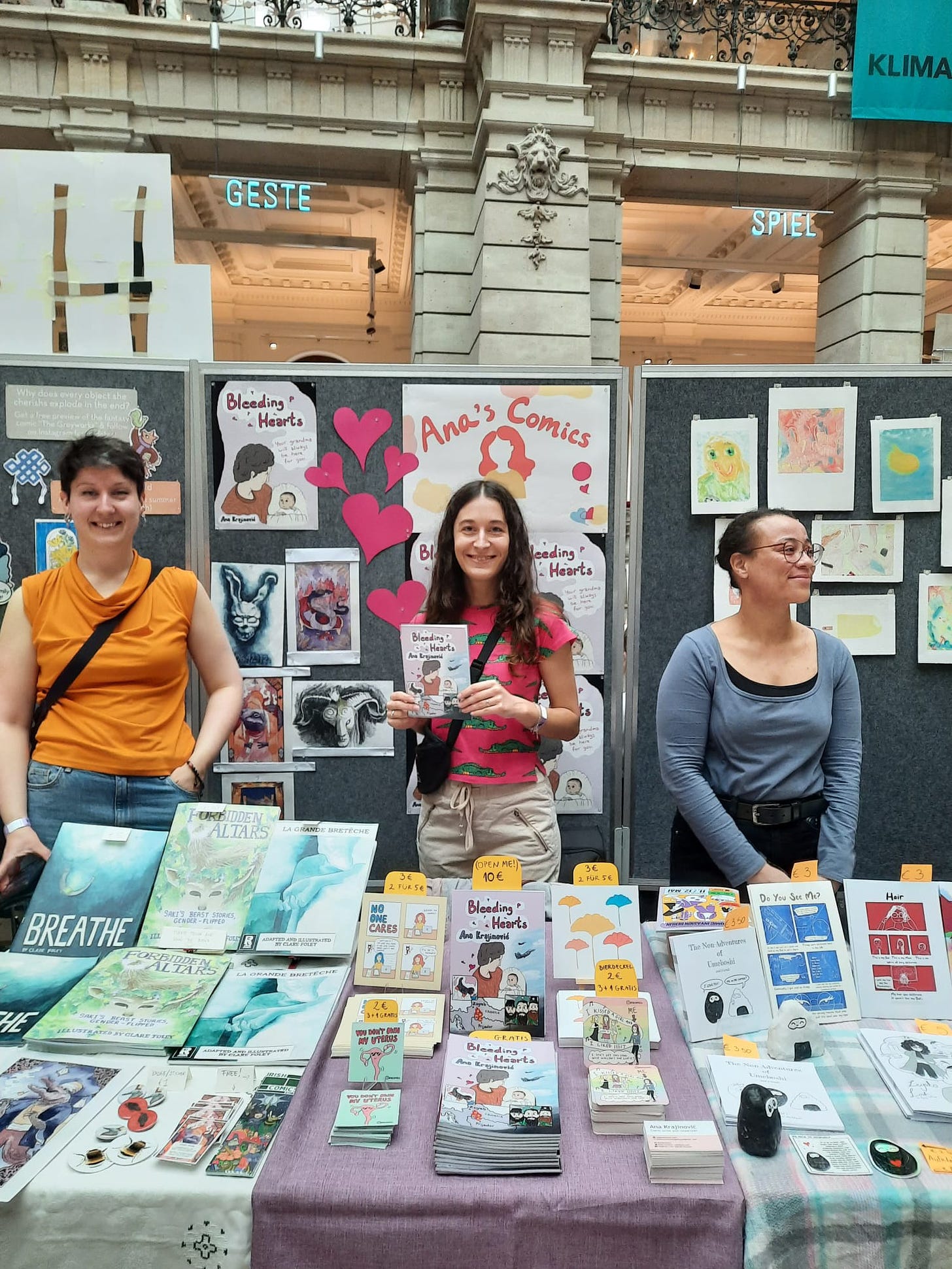 The photo shows a comic convention with three participants standing behind their tables. I am in the middle holding my comic "Bleeding hearts".