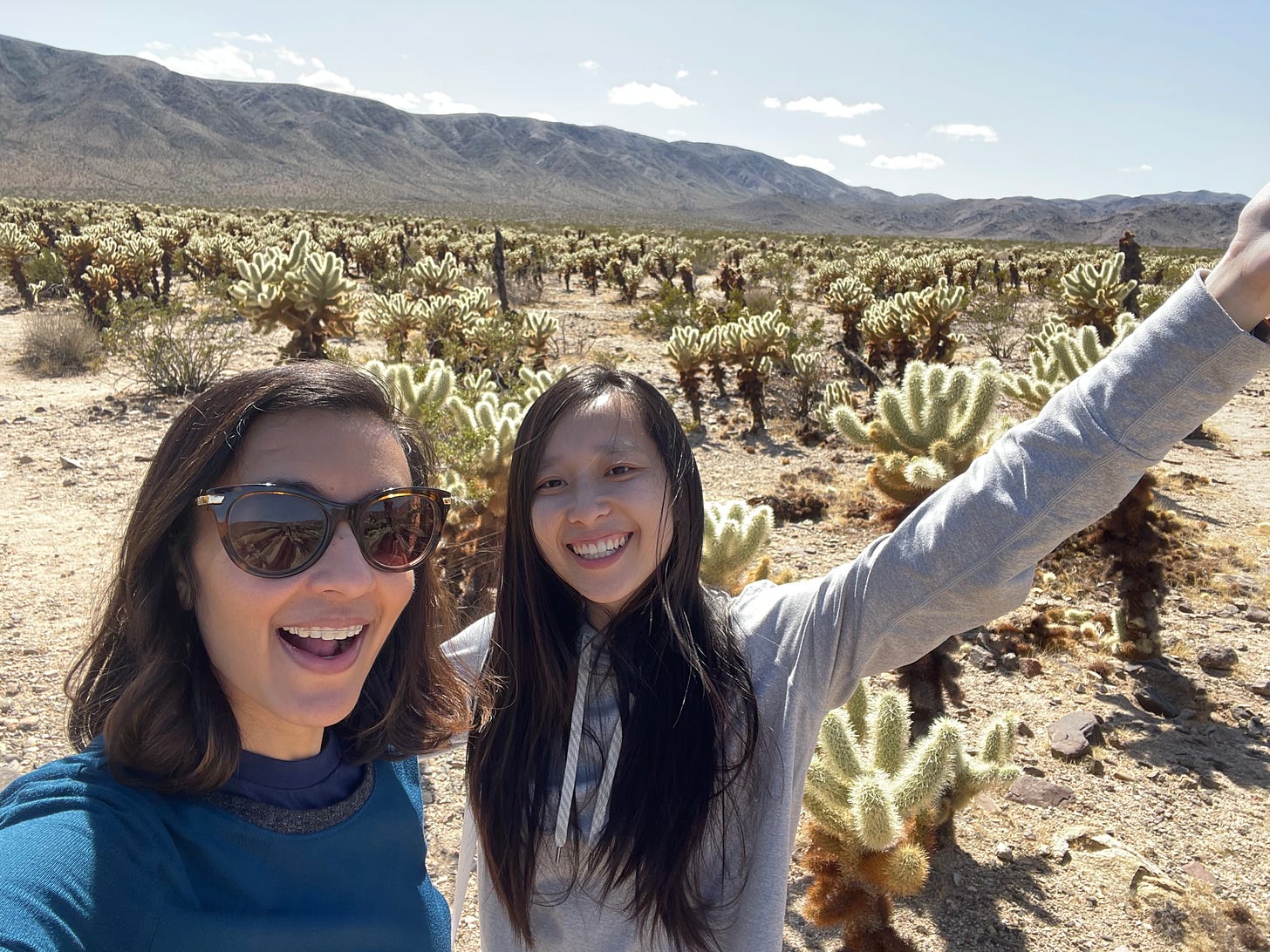 Two woman stand in front of a cactus garden in the desert. The woman on the right has long black hair and wears a gray jacket. The woman on the left has short dark brown hair, sunglasses, and a blue shirt.