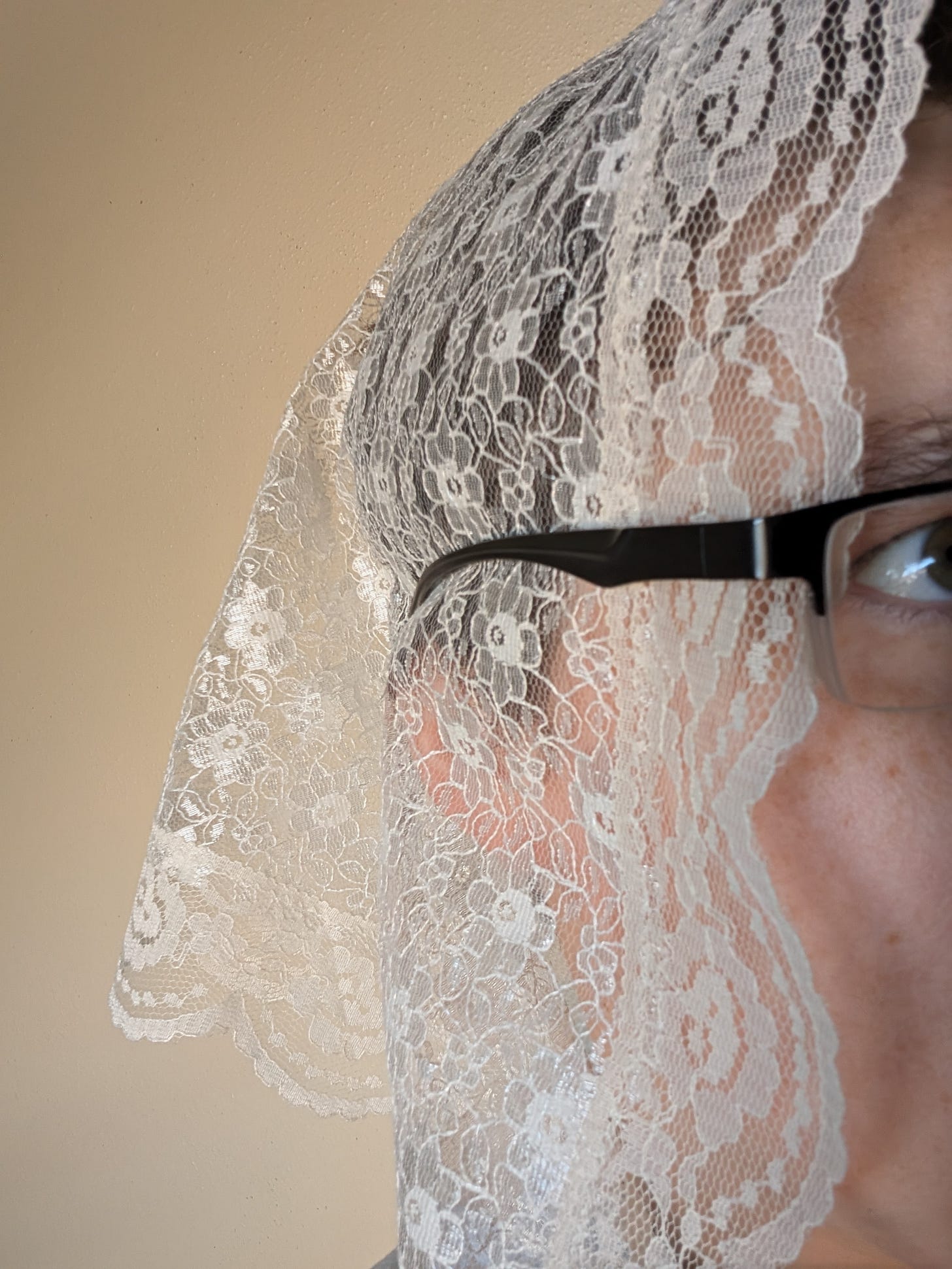 A woman wearing a long lace headcovering and glasses. The temples of the glasses are worn on the outside of the lace, holding it to the head.