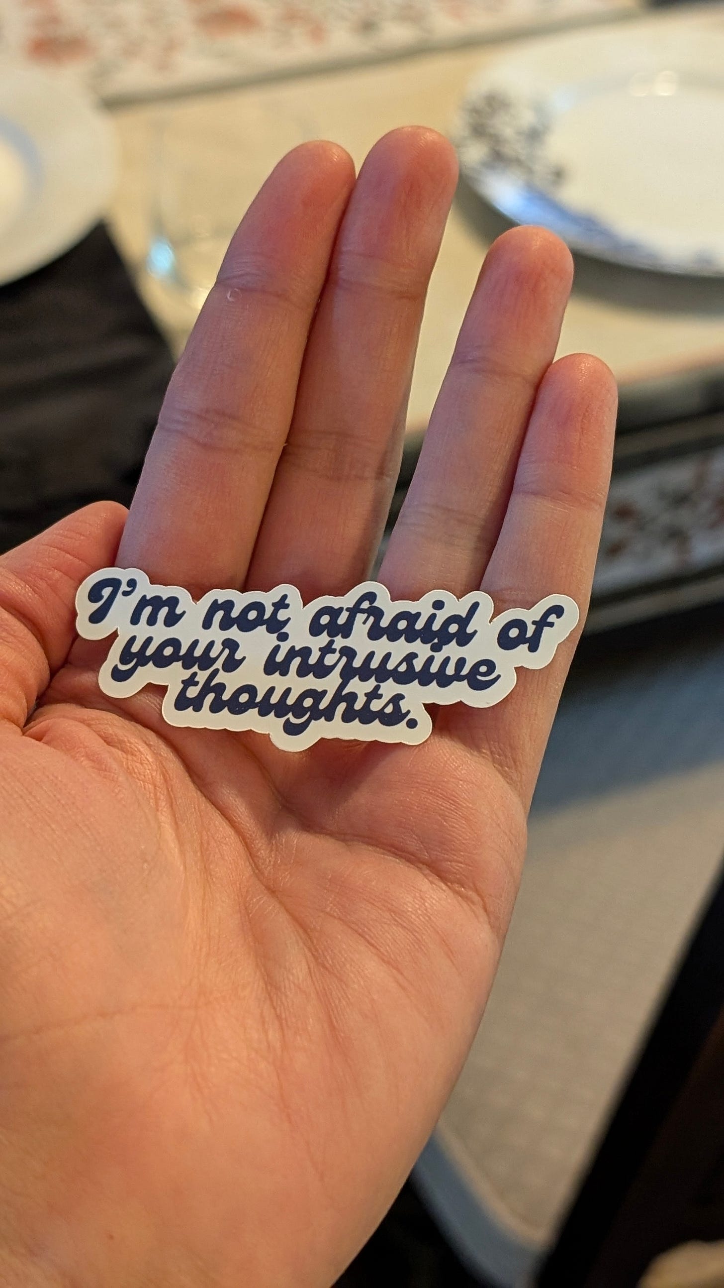 My hand holding a sticker that, in fun vintage font, says "I'm not afraid of your intrusive thoughts."