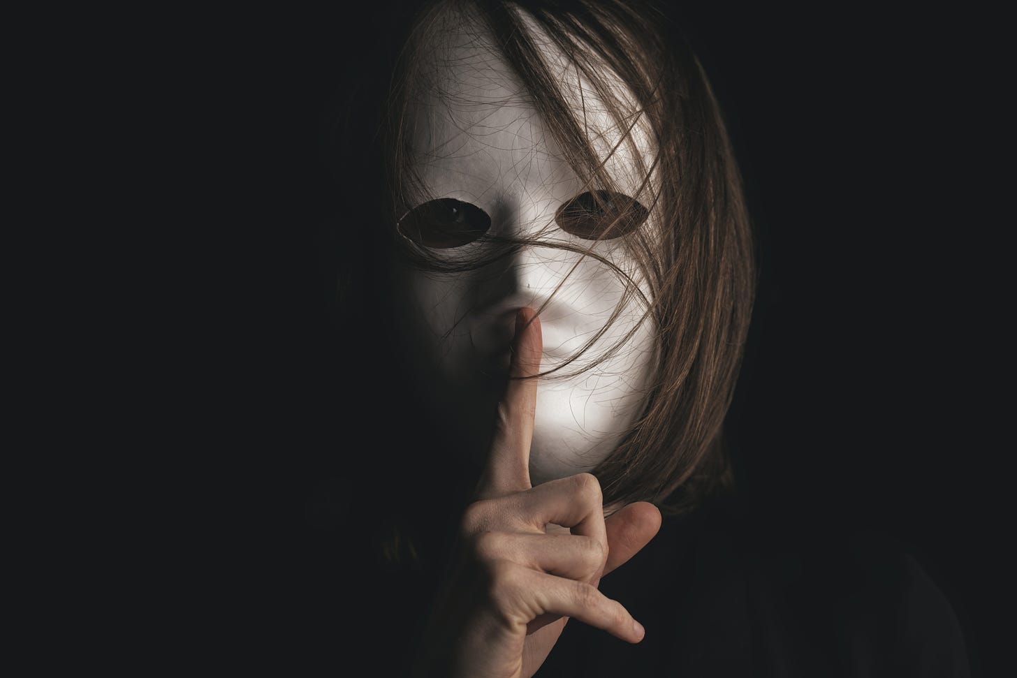 a figure in a white mask with medium length brown hair appears out of the darkness, holding their finger over their mouth in a shhh motion