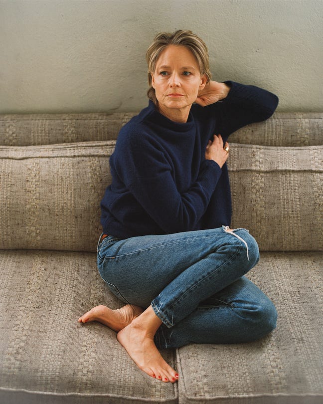 A portrait photograph of the actor Jodie Foster curled up barefoot on a couch, wearing jeans and a navy-blue sweater.