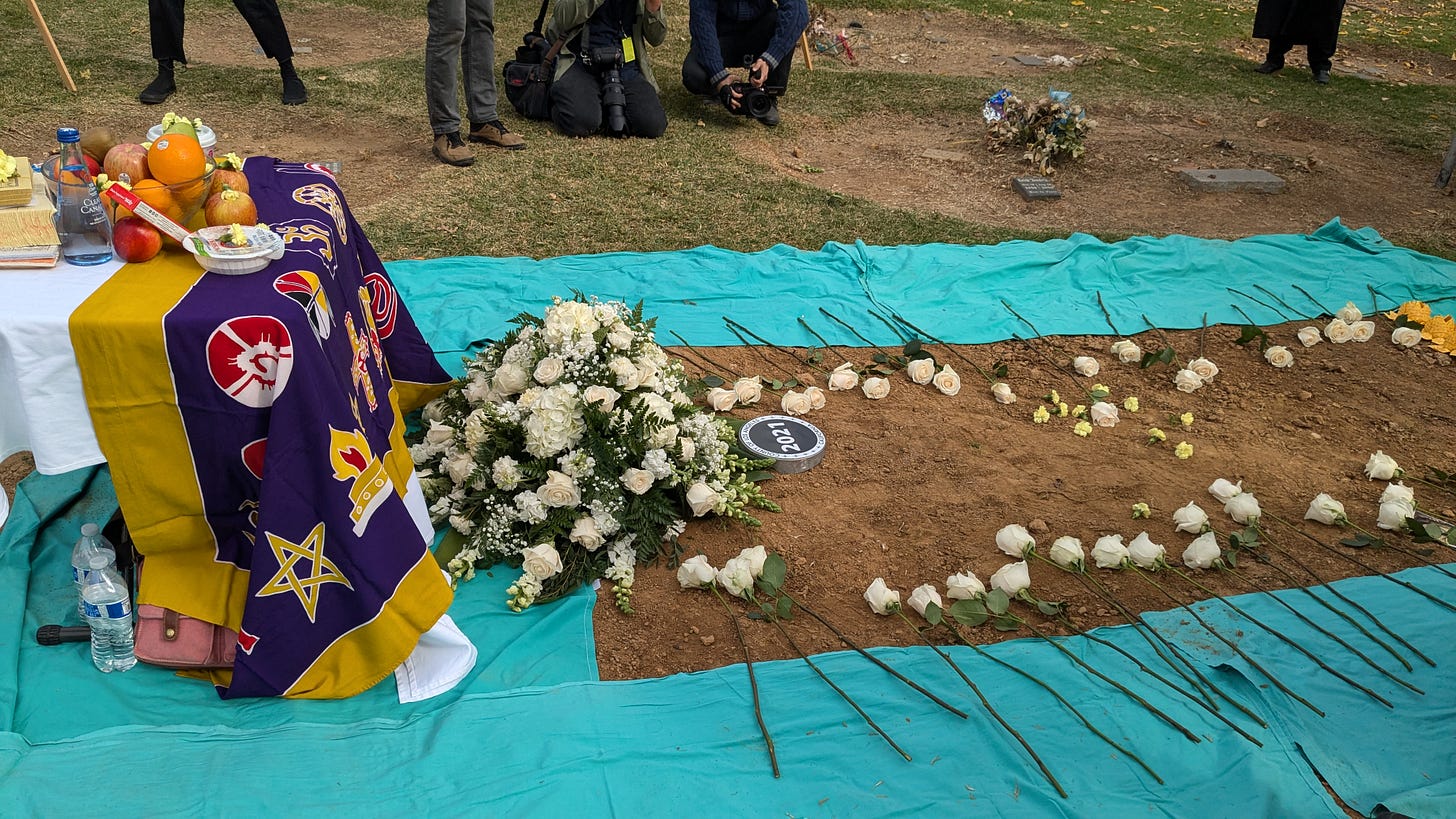 Offerings of fruit sit on a table covered with a ceremonial cloth at the head of a freshly dug mass grave. White flowers lie on the grave, which is labeled 2021. Green tarps surround the grave