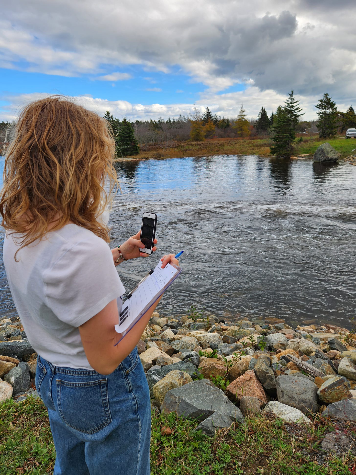 A young woman wearing a white tee shirt and blue jeans standing on a rocky shore is taking a picture of the water with her iphone with her left hand. She holds a pen and papers on a clipboard in her right hand.