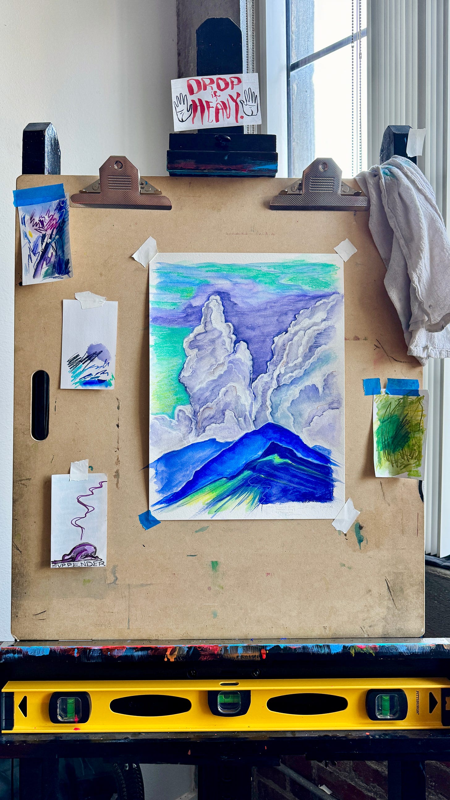 colorful watercolor of clouds sky and mountains in hues of purple green and blue taped to an artboard on an easel.  A handwritten note reads "DROP IF HEAVY".