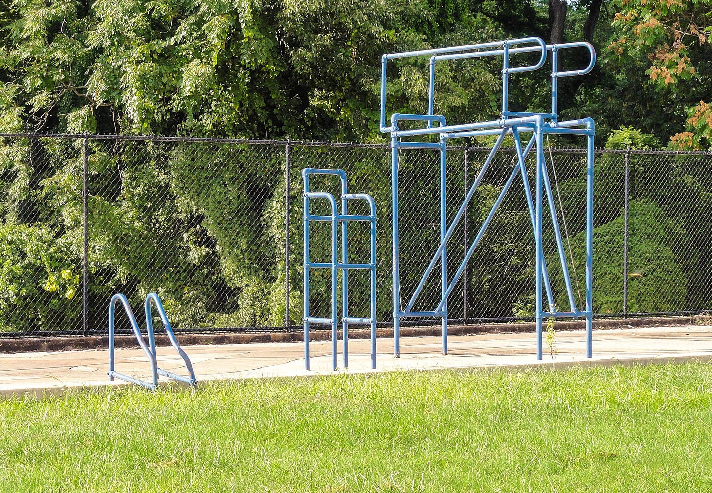 This photograph shows the blue metal-structured pool ladder, lifeguard chair, and diving board on the side of the Druid Hill Pool No. 2 memorial. The inside of the pool is filled with soil and grass.