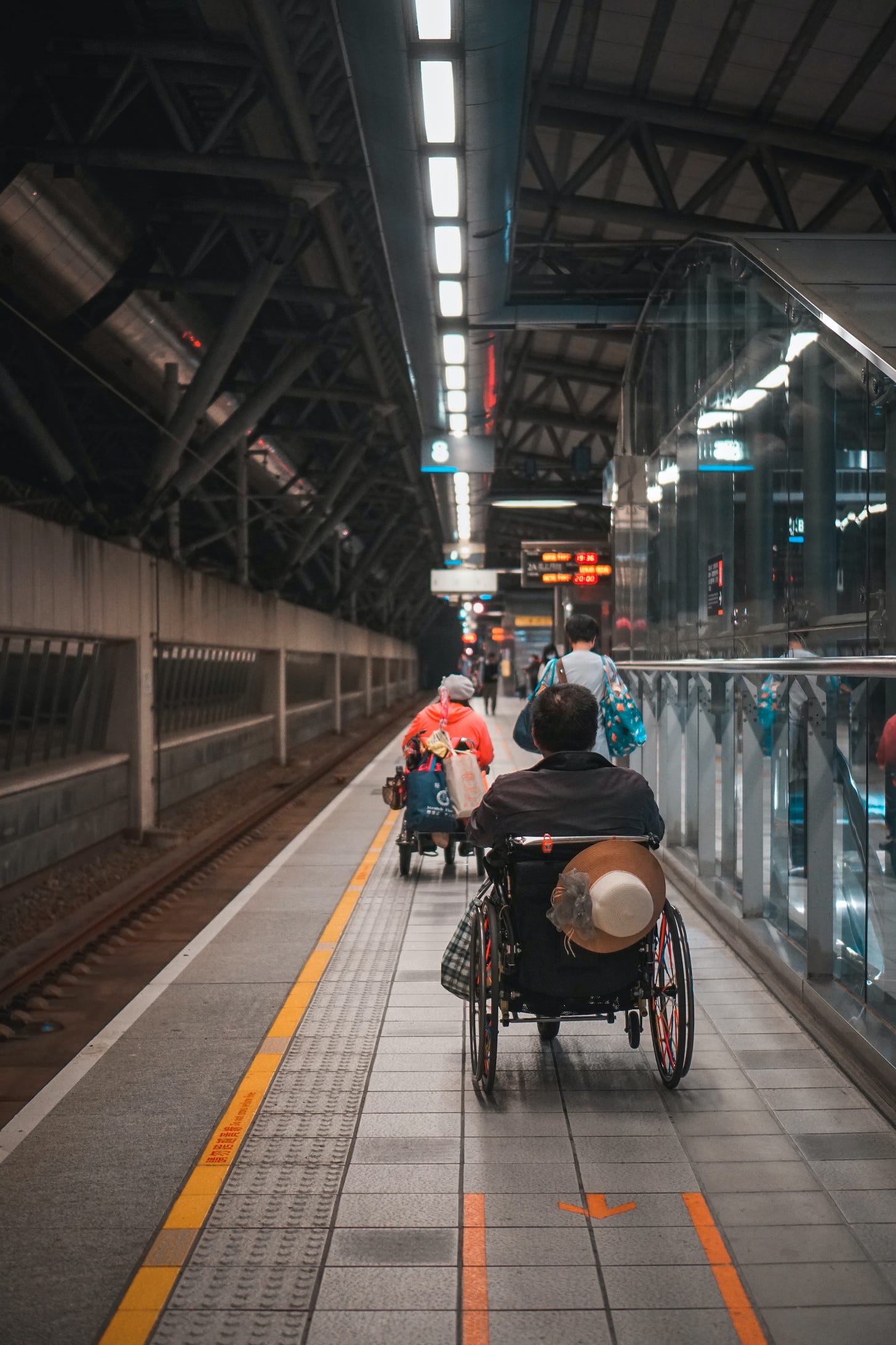 Picture of a train platform with a couple of people in wheelchairs with their backs to the camera making their way down the platform .